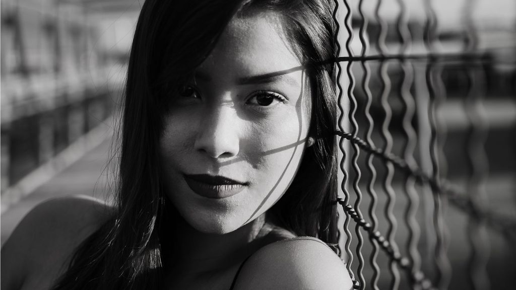 A black and white photo of a sexy woman smirking with a chain fence behind her.