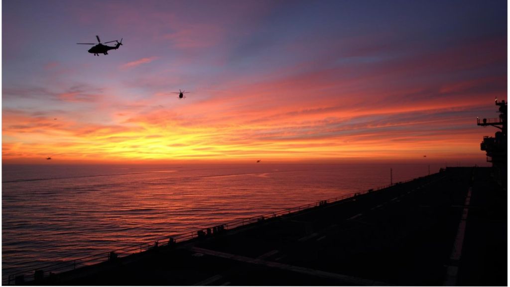 Silhouetted helicopters fly above a calm ocean at sunset beside the dark outline of an aircraft carrier deck, evoking a tense, reflective mood that symbolizes the quiet war within.AI generated image.